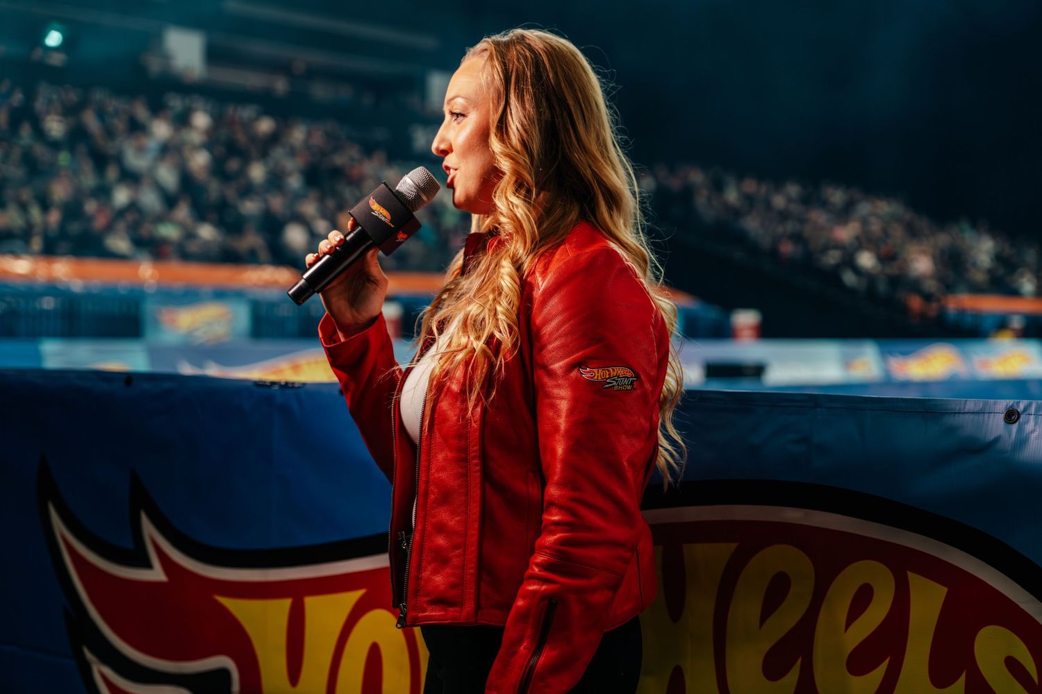 A blonde woman in a red jacket sings into a microphone at a Hot Wheels live event.