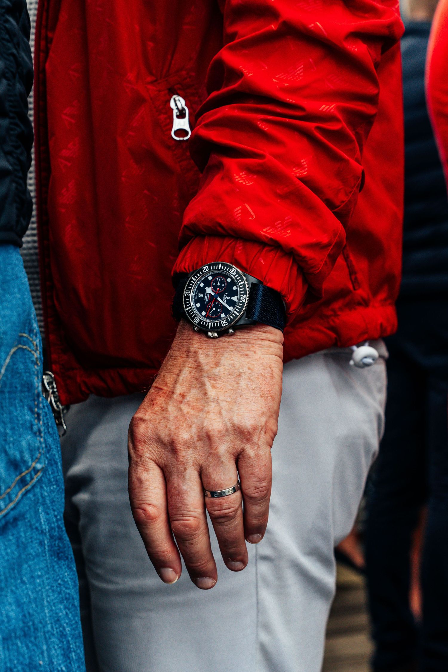 Close-up of a man's hand wearing a black chronograph watch and ring with a red jacket.