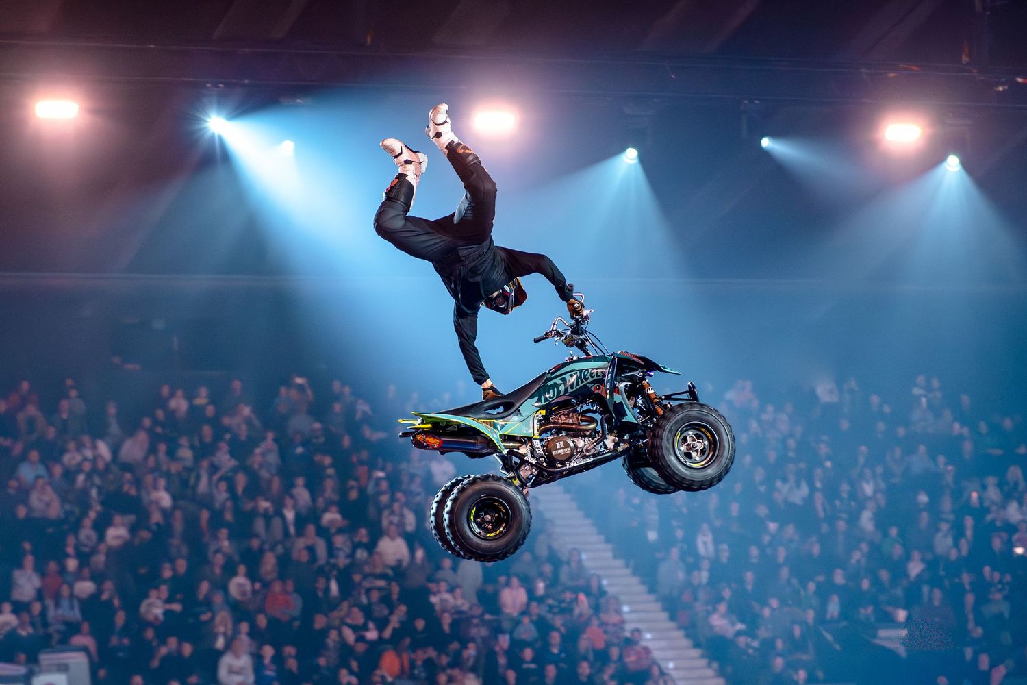 A fearless stunt performer stands on a quad bike mid-air before a cheering arena crowd.