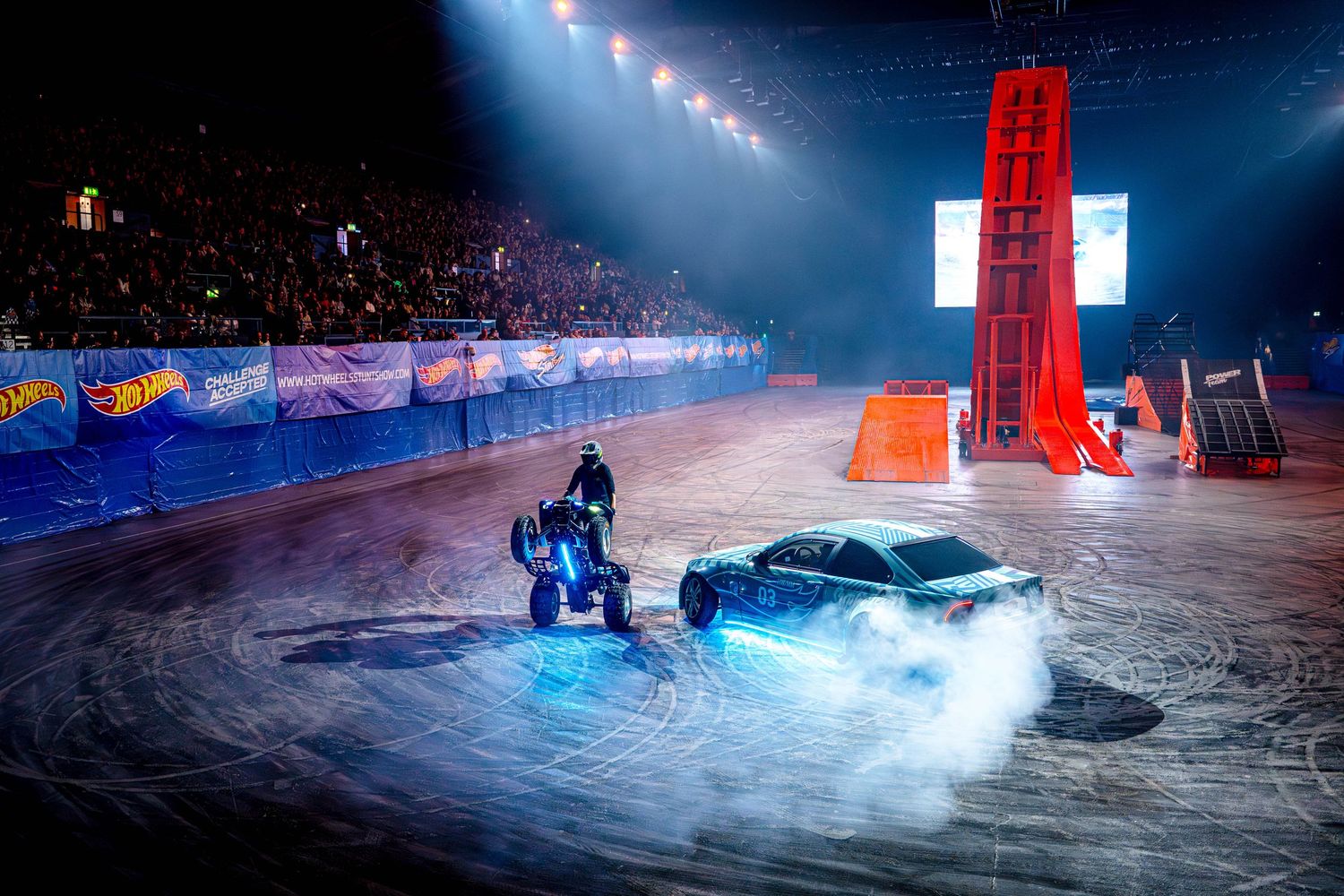 Hot Wheels live stunt show featuring a car and motorcycle performing on an indoor arena floor.