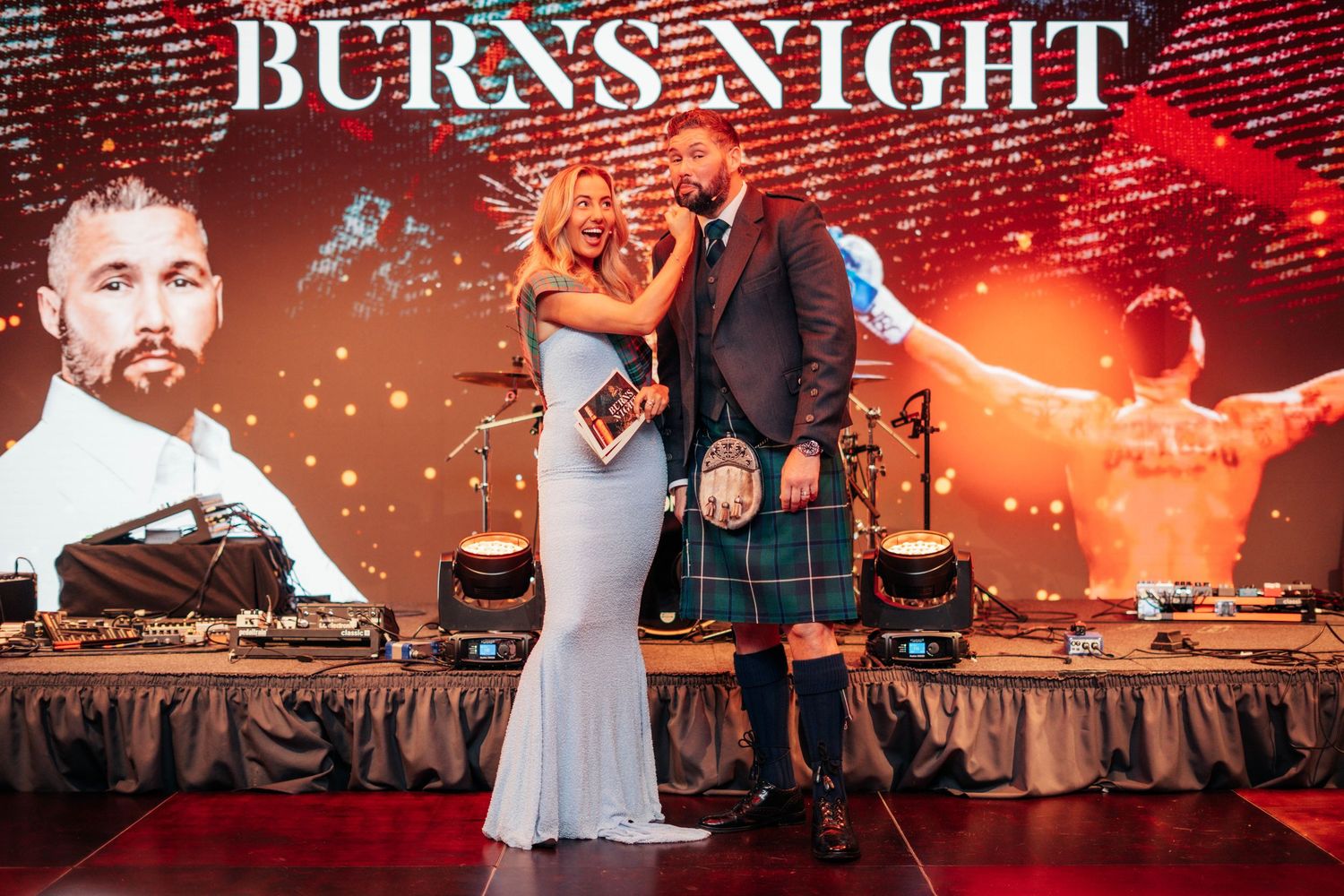 A couple poses on stage at a Burns Night event with a large illuminated backdrop behind them.