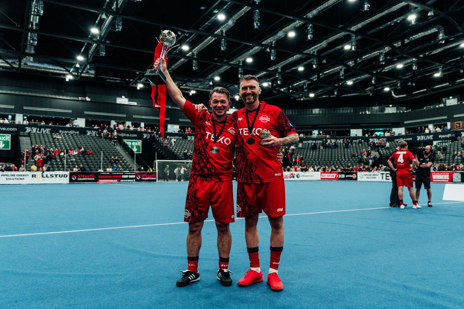 Two athletes in red uniforms celebrating with trophy in indoor sports arena.