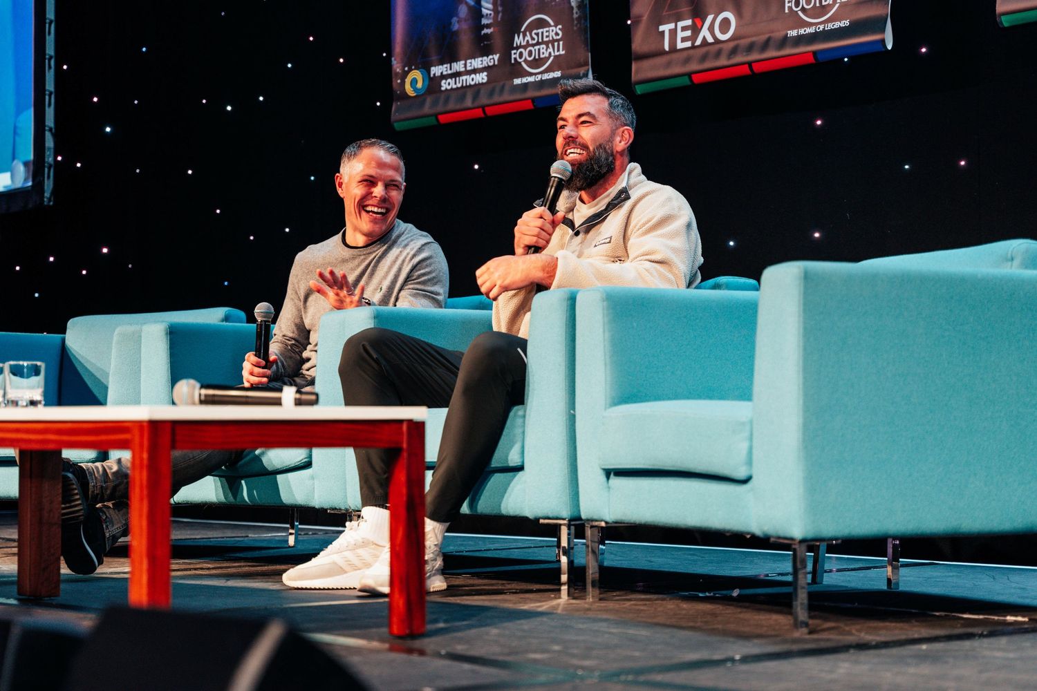Two men laughing on stage seated in blue chairs at conference with microphones.