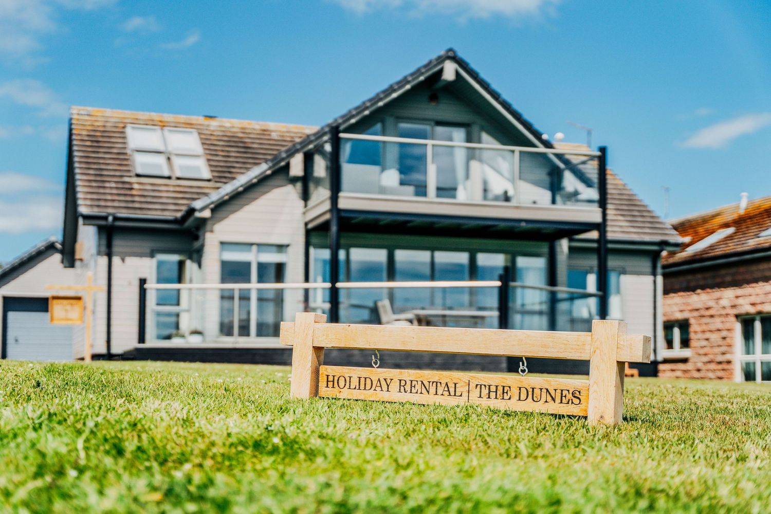 Holiday rental sign for The Dunes in foreground with coastal property and blue sky behind.