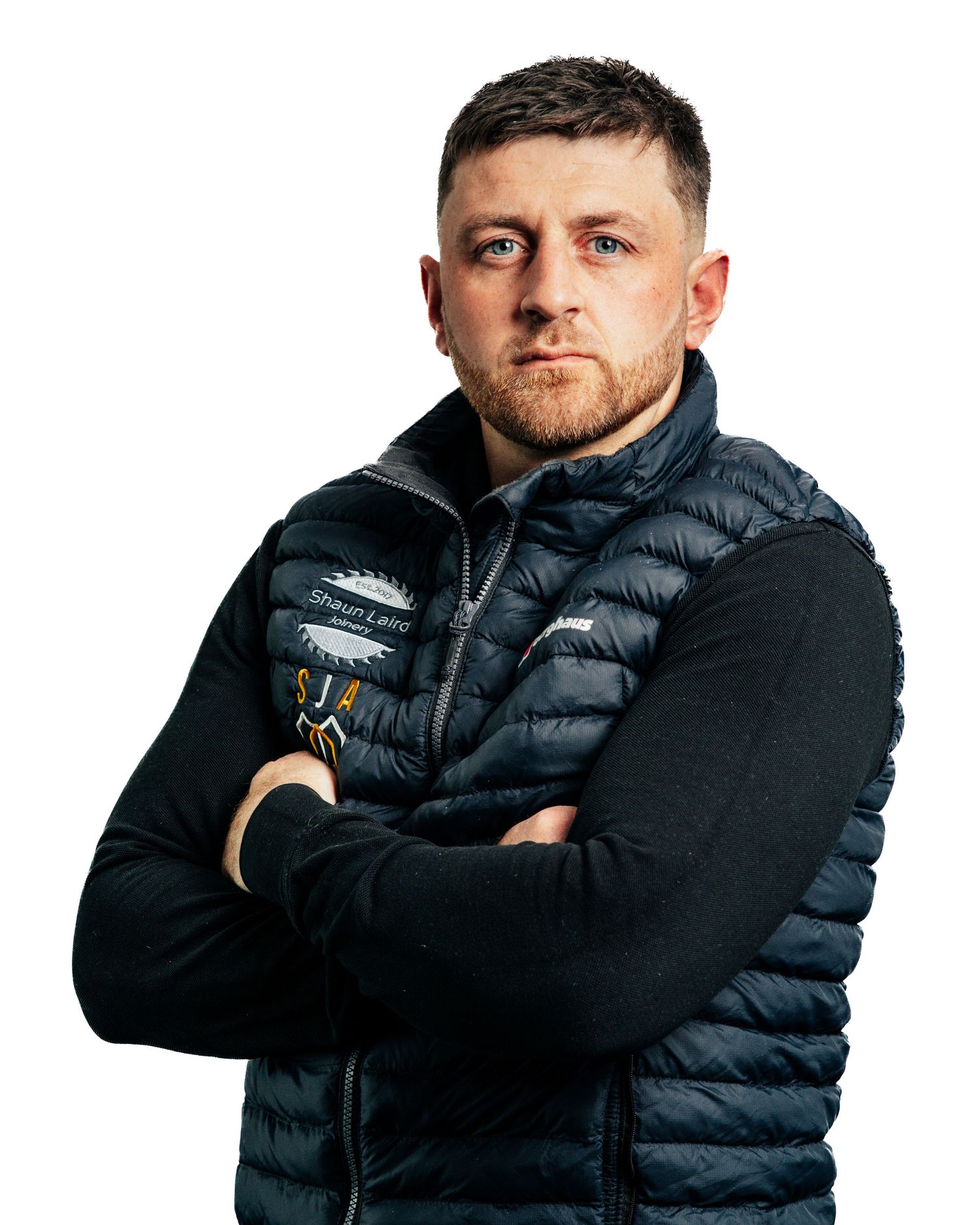 Serious man in black puffer vest with arms crossed against white background.