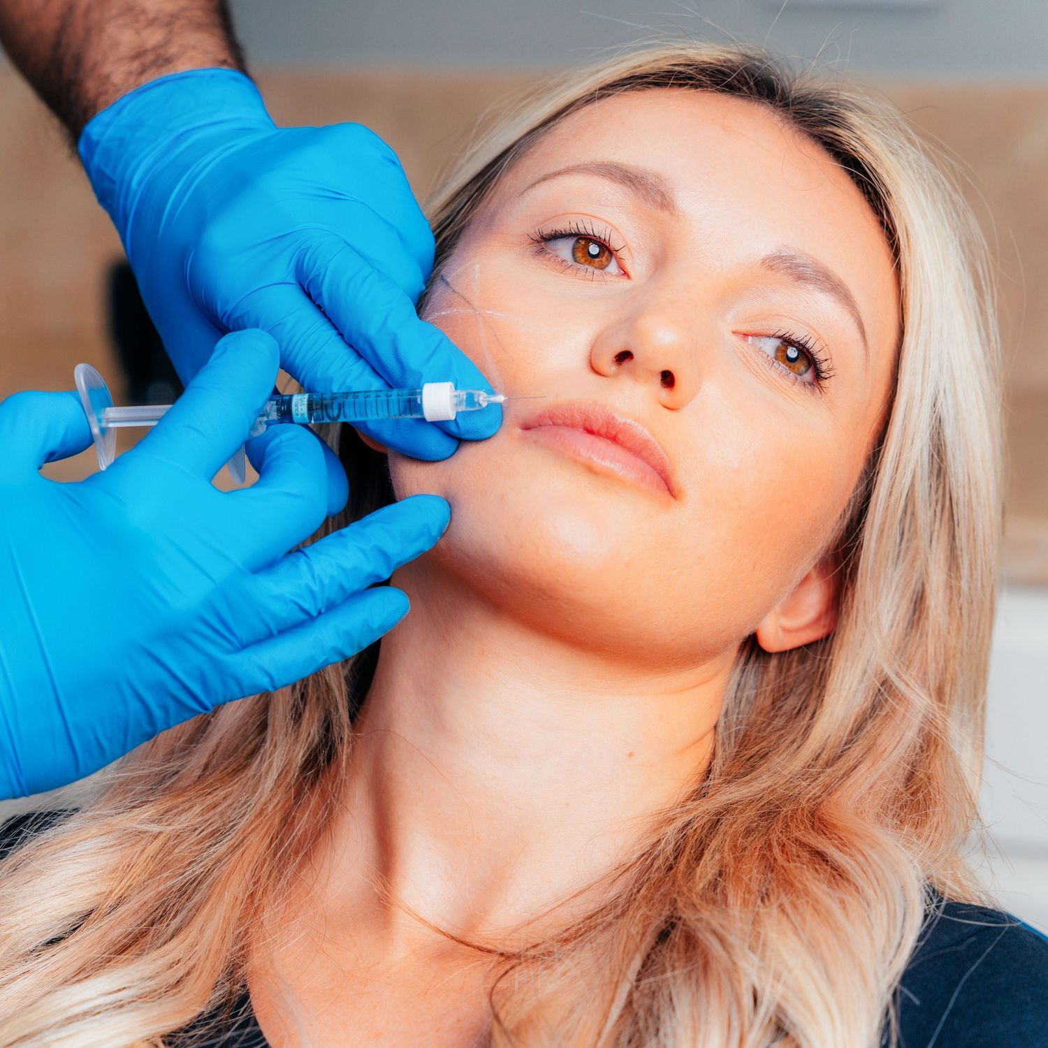 A medical professional in blue gloves administering a facial filler injection to a blonde woman.