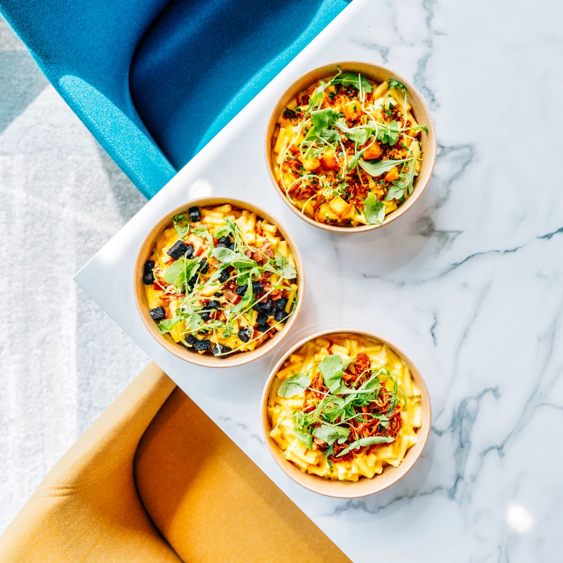 Three mac and cheese bowls topped with crispy garnishes and herbs on a marble surface.