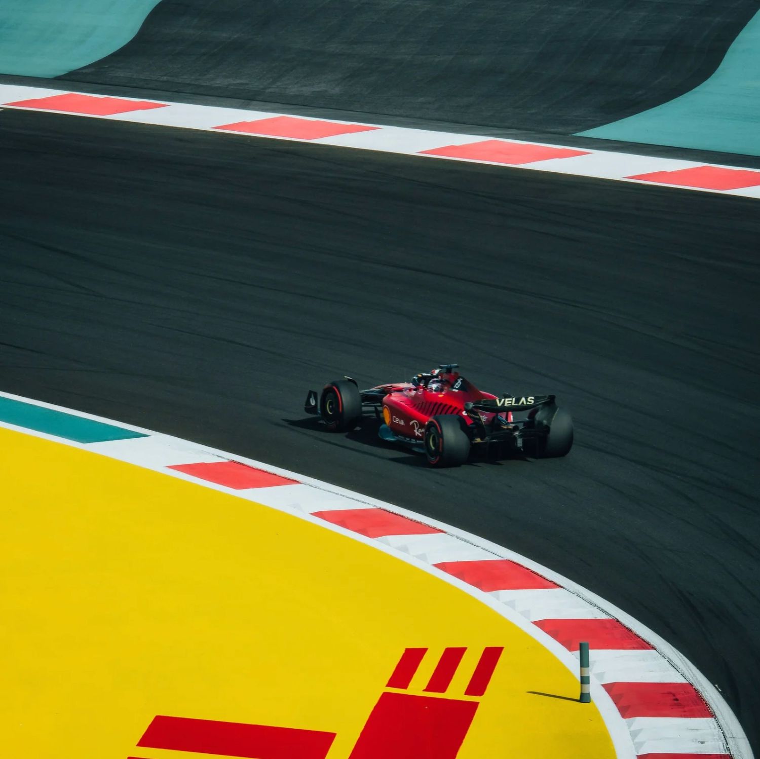 A red Ferrari Formula 1 car races through a colorful corner at a sunny circuit with yellow and red curbs.