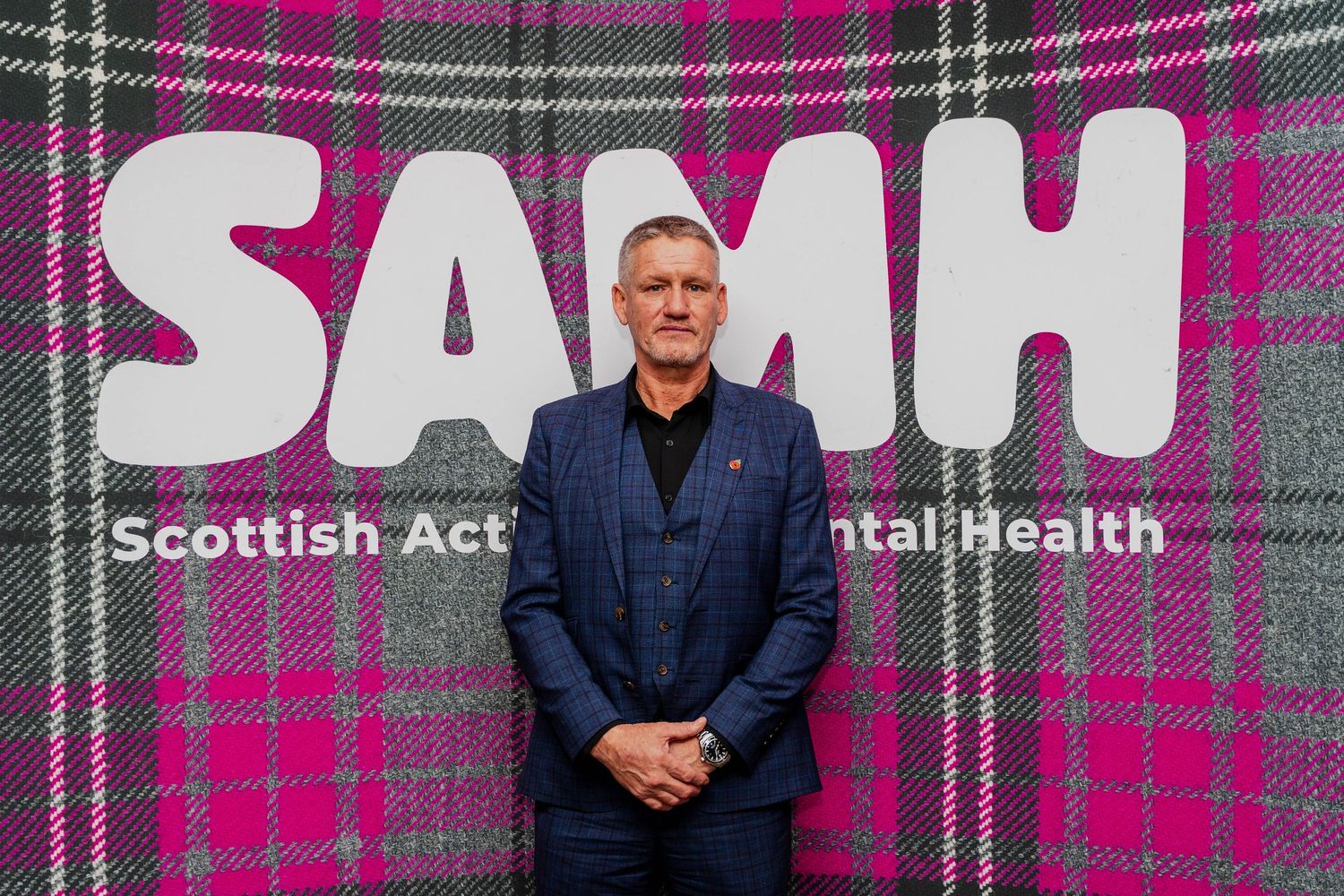 Man in blue suit standing before SAMH Scottish Action for Mental Health pink tartan banner.