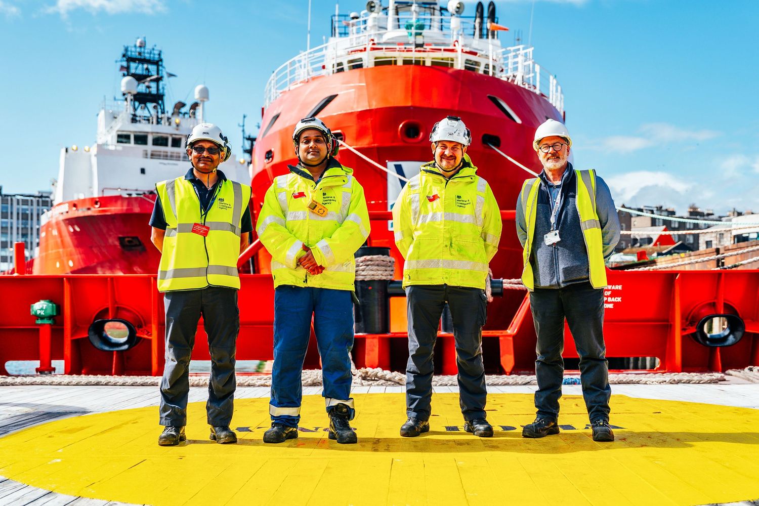 Four crew members in yellow high-visibility jackets pose together on the deck of a red offshore supply vessel.