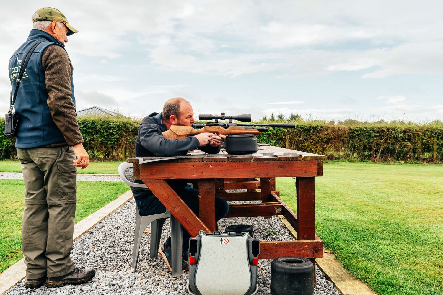 A man aims a scoped rifle at an outdoor shooting range while an instructor observes nearby.