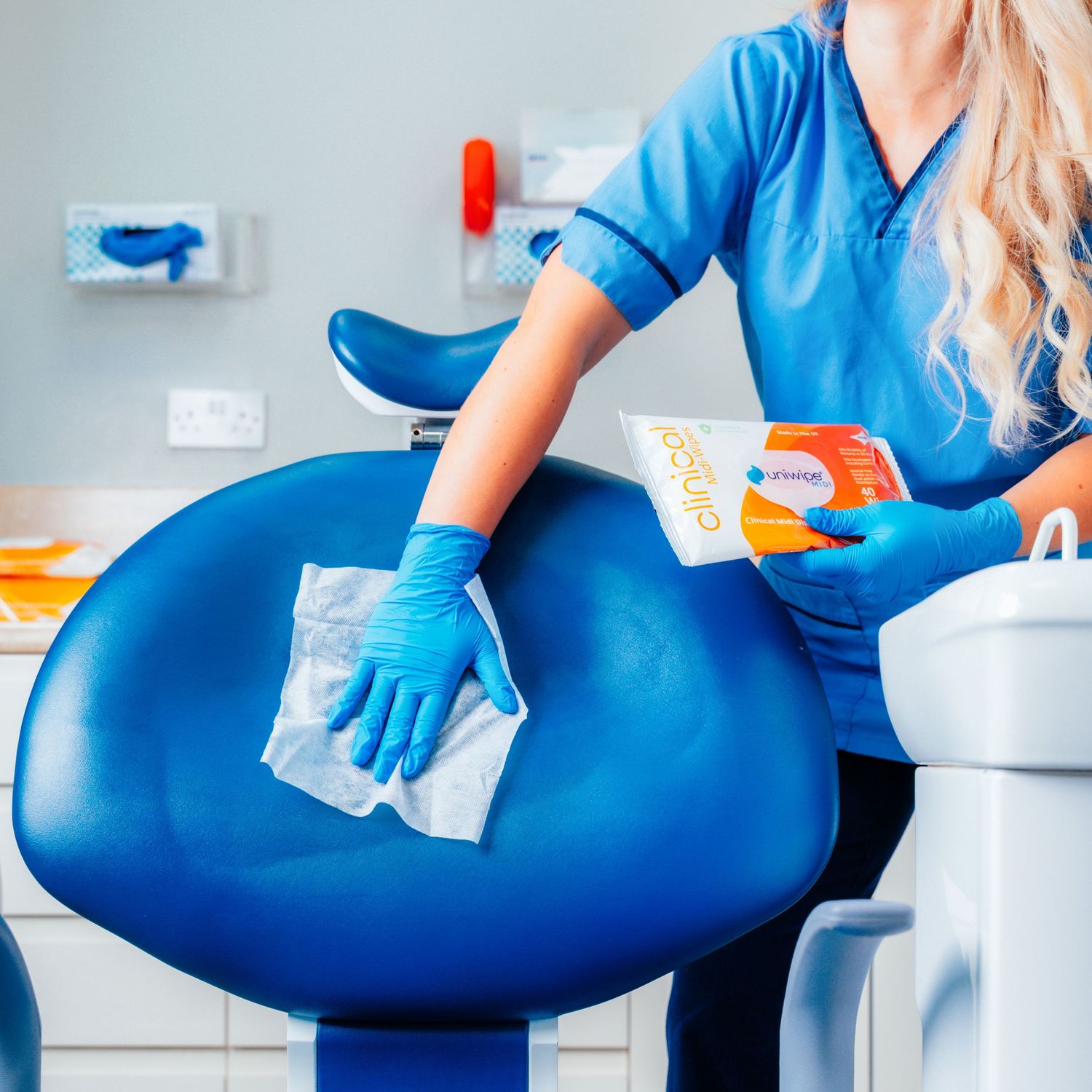 Blonde nurse in blue scrubs cleaning a dental chair with Uniwipe disinfectant wipes in a clinical setting.