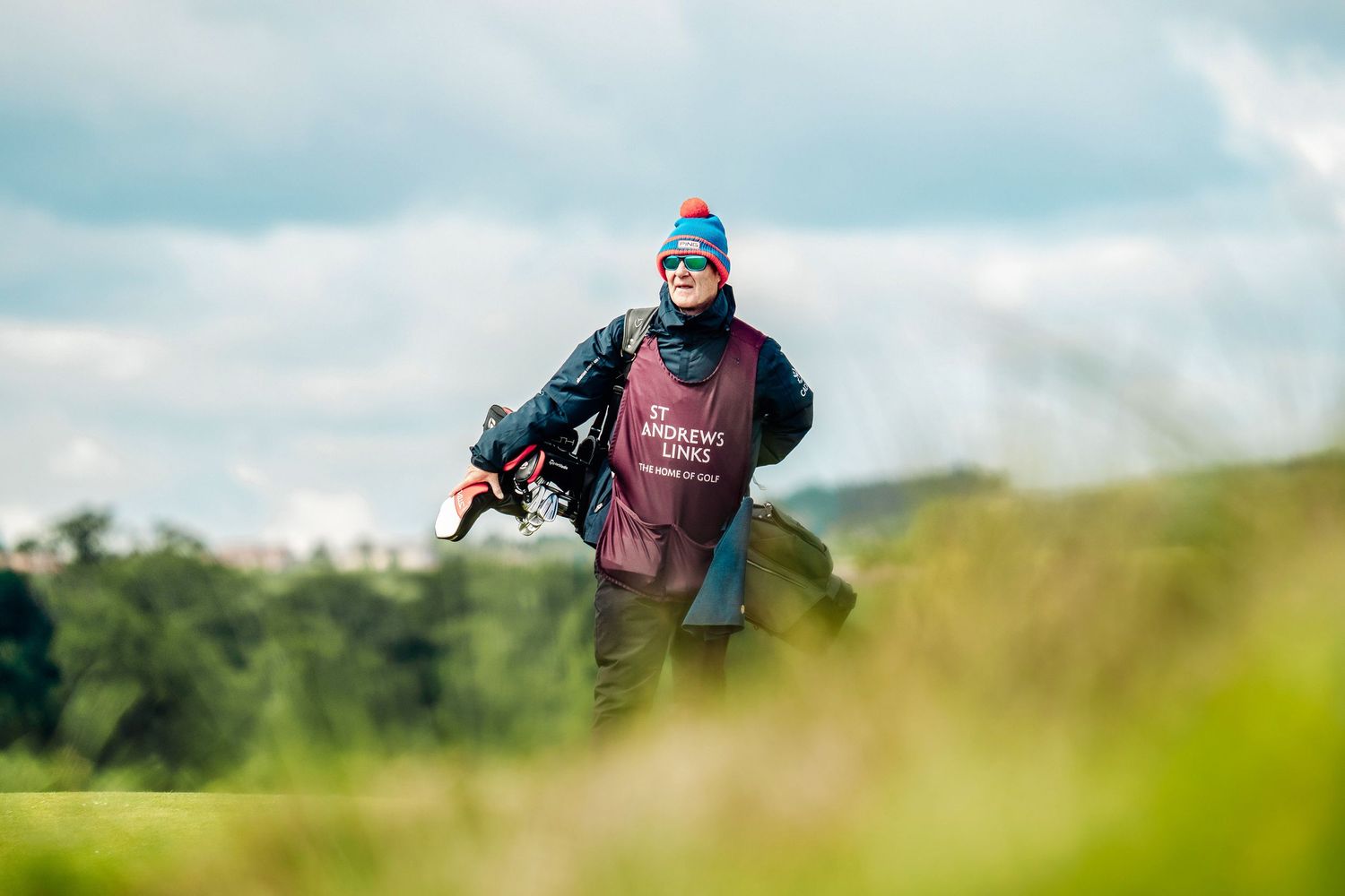 A caddie carrying a golf bag walks across a lush green golf course wearing a tournament bib and colorful hat.