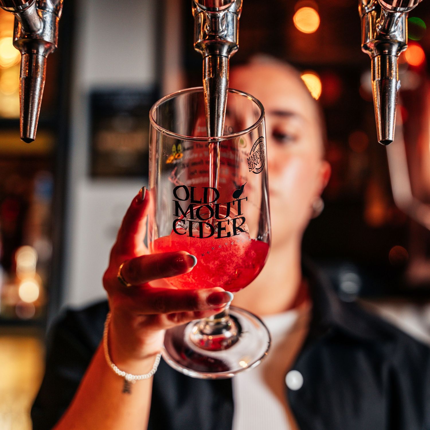 Bartender holding a branded Old Mout Cider glass filled with bright red cider at a bar.