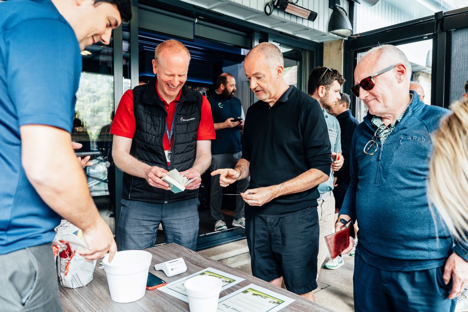 Group of men socializing at an outdoor event table with drinks and materials.