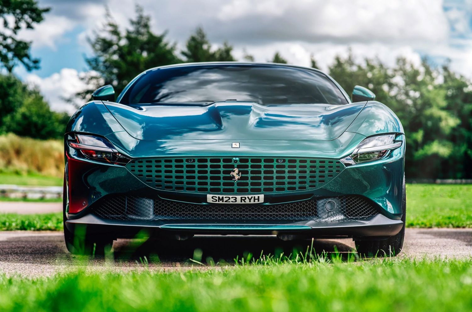 Dark green Ferrari Roma sports car front view on a scenic driveway with lush grass and cloudy sky.