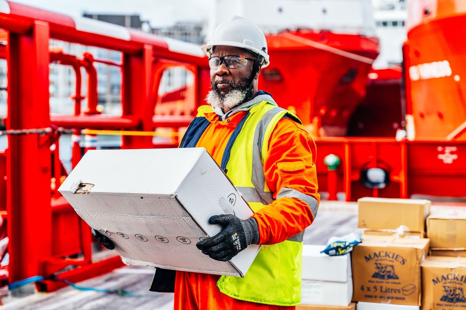 Offshore worker in orange coveralls and hi-vis vest carries a box on a supply vessel.