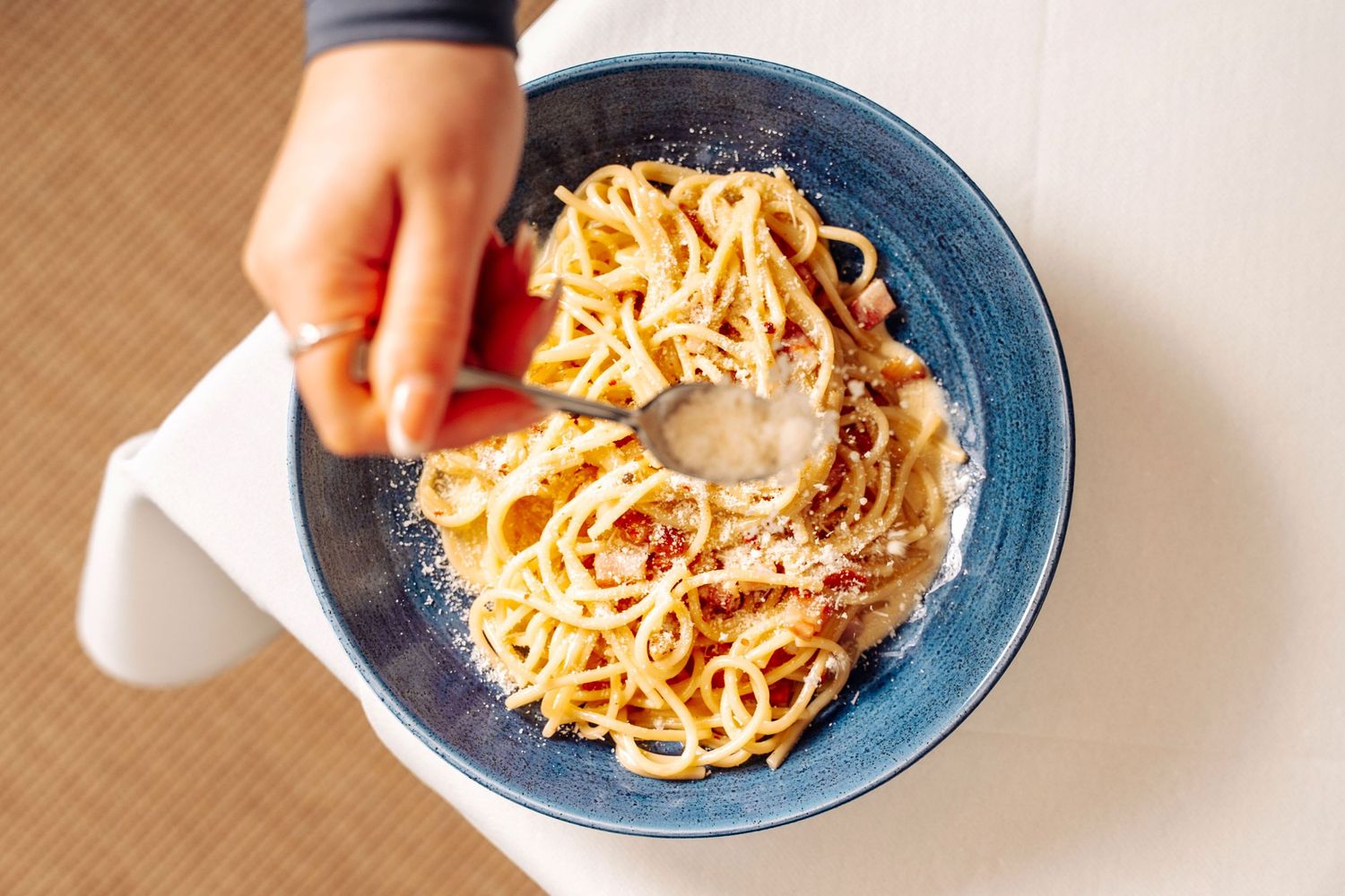 Hand twirling spaghetti carbonara with a spoon in a blue bowl, topped with parmesan cheese.