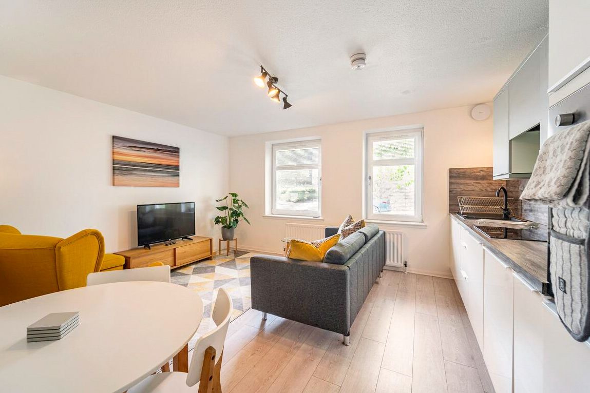 Bright modern living room with grey sofa, mustard chairs, light wood floors, white kitchen, and large windows.