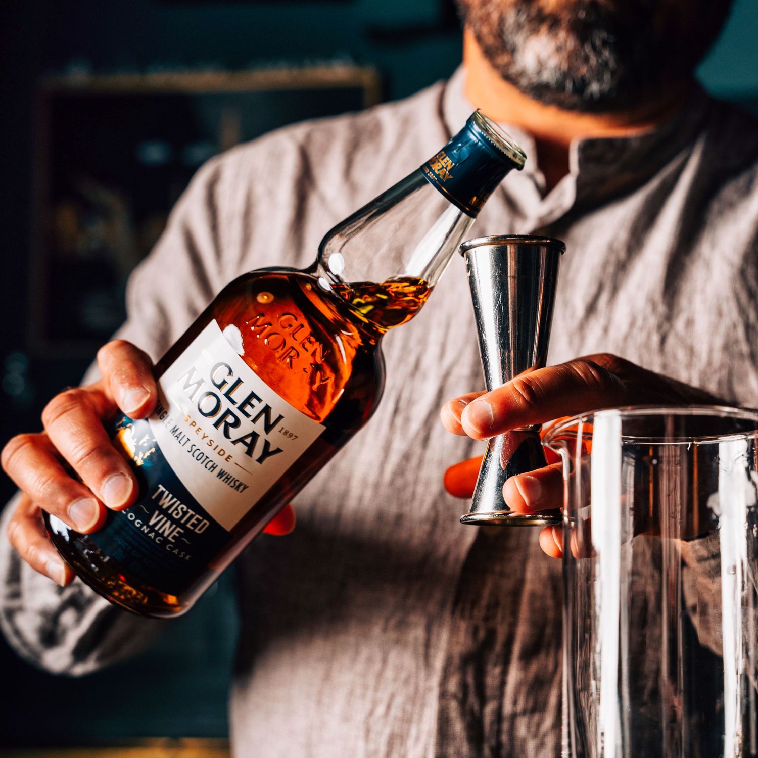A bartender pouring Glen Moray whisky into a silver jigger at a bar counter.
