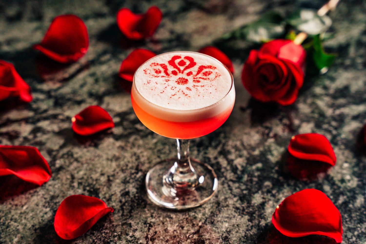 A pink frothy cocktail in a coupe glass with a floral design, surrounded by red roses and petals.