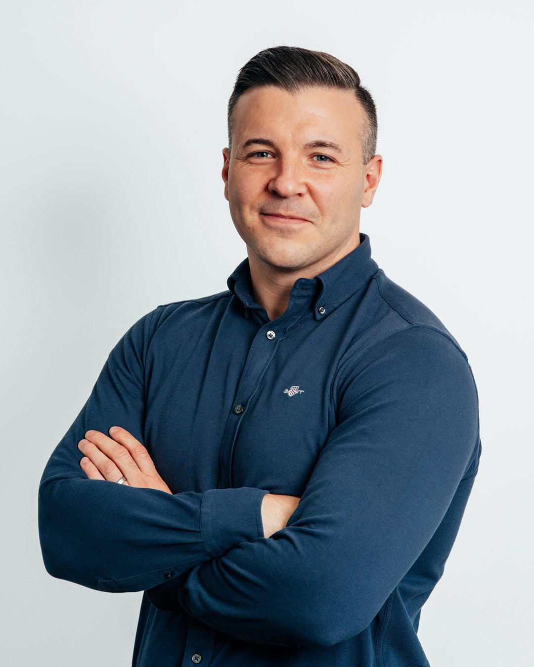 Professional man in navy blue shirt standing with arms crossed smiling against light background.