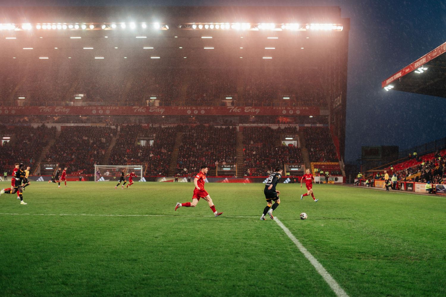 Football players in action on a rain-soaked pitch under bright floodlights at a red-themed stadium.