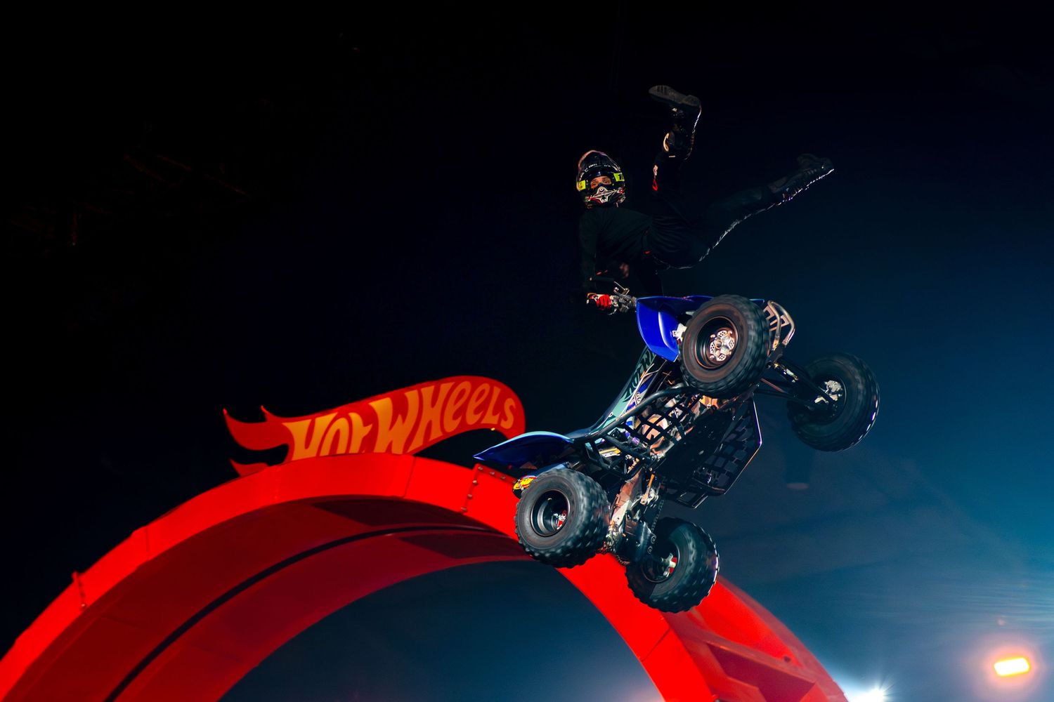 A quad bike rider performs a breathtaking aerial stunt over a Hot Wheels branded ramp at night.