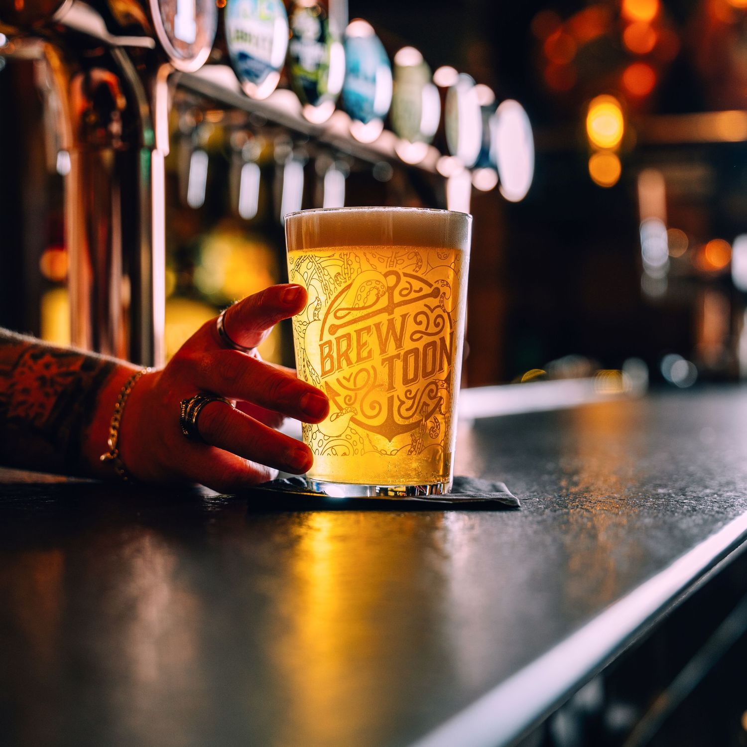 Hand holding a branded Brew Toon pint glass of golden lager at a bar with tap handles.