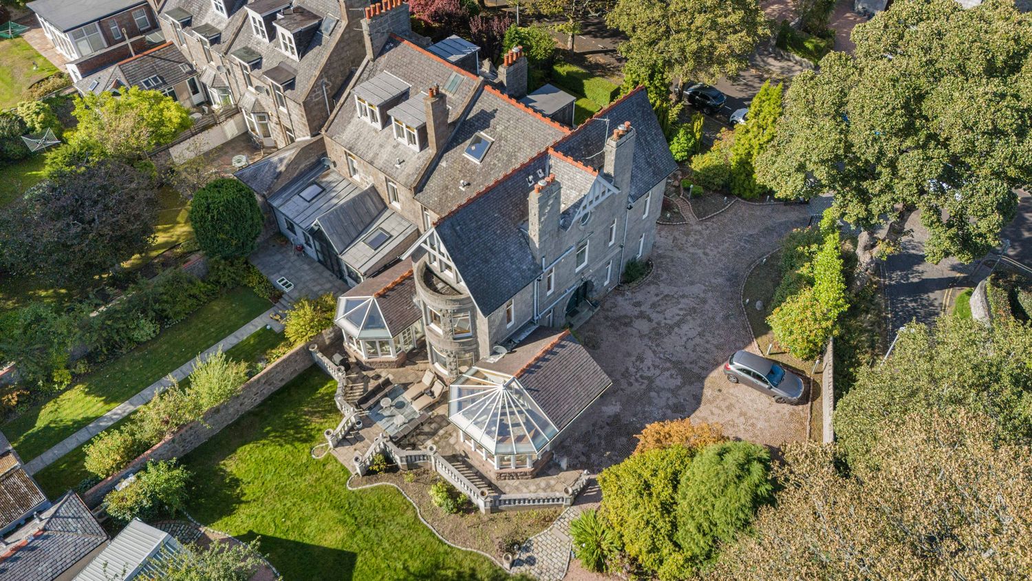 Aerial view of a large stone manor house with conservatory extension surrounded by mature trees and gardens.