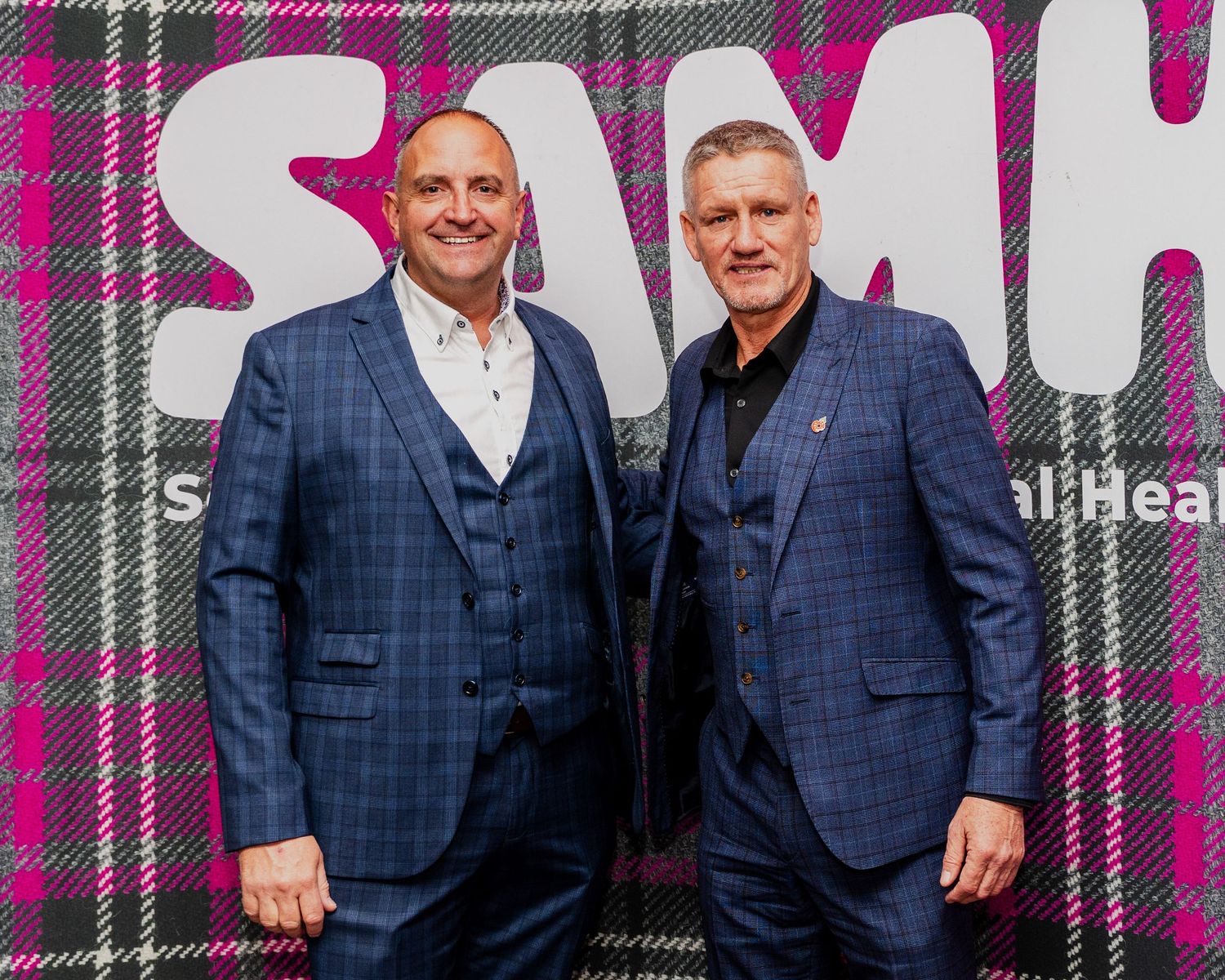 Two men in blue suits posing together in front of SAMH mental health branded backdrop.