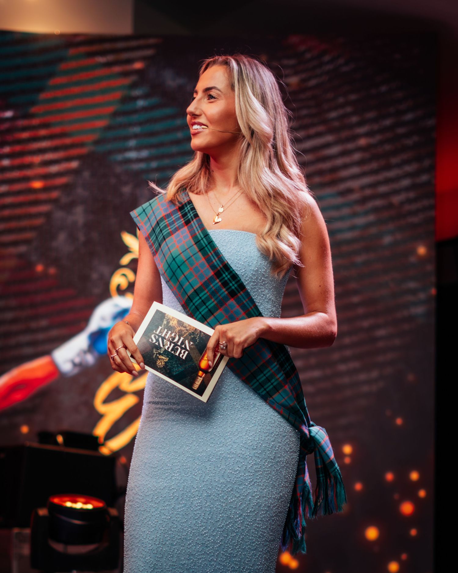 Elegant woman in light blue dress with tartan sash holding a program at a Burns Night event.