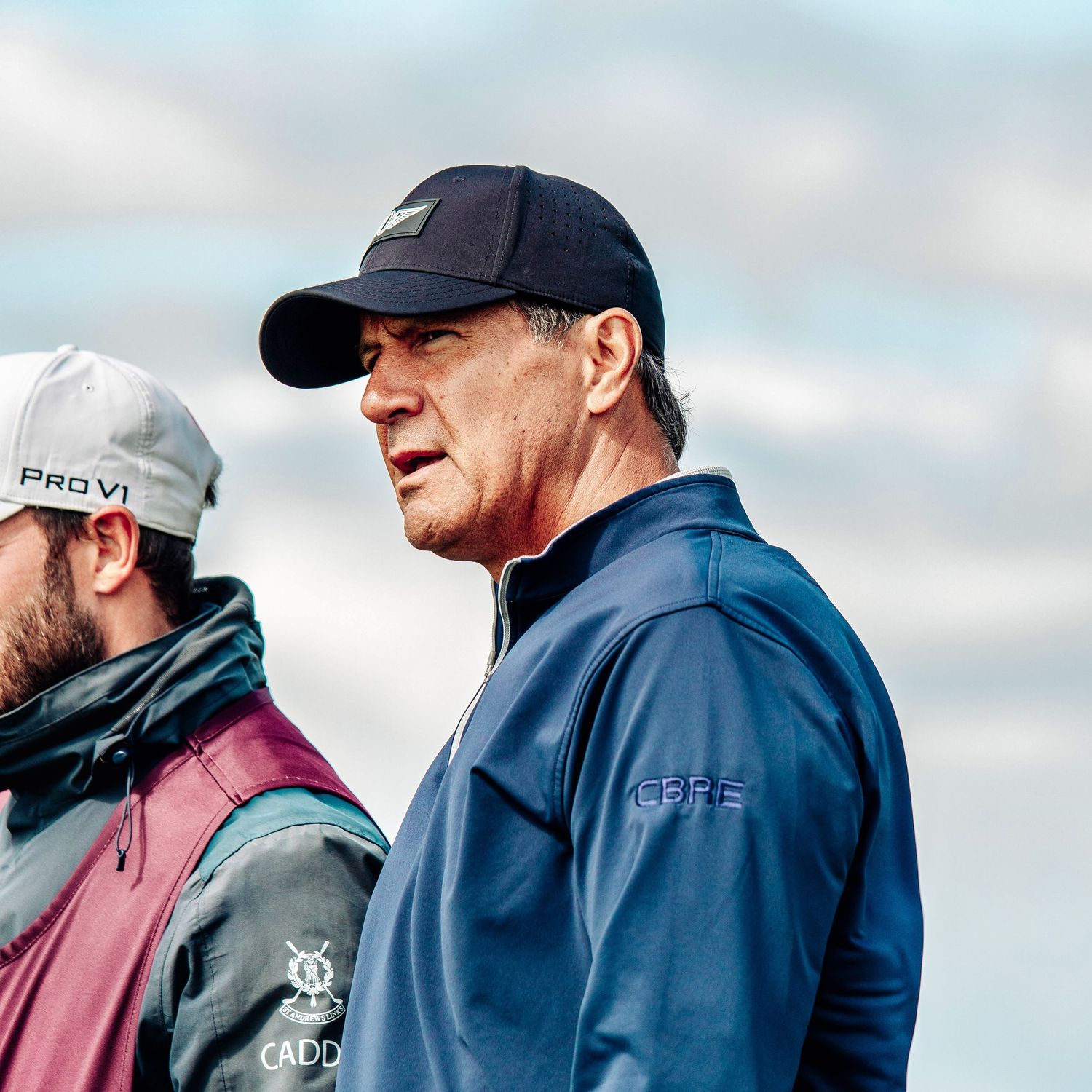 Two men on a golf course, one wearing a navy cap and blue jacket with CBRE logo, against a cloudy sky.