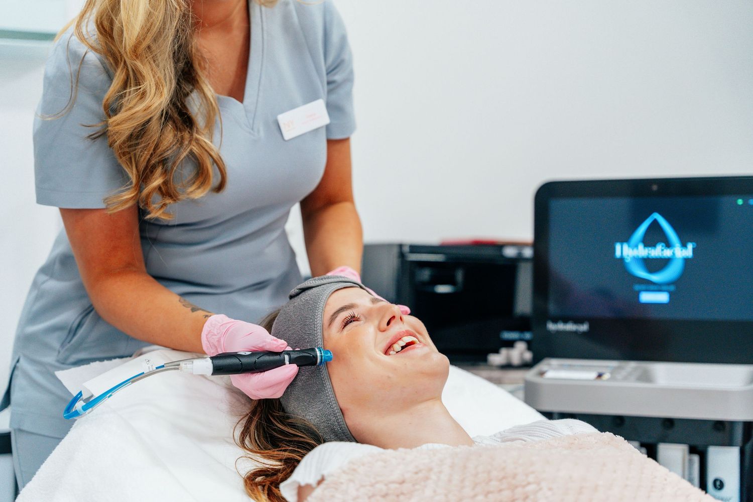 Smiling woman receiving a HydraFacial treatment from a blonde aesthetician in a professional beauty clinic.