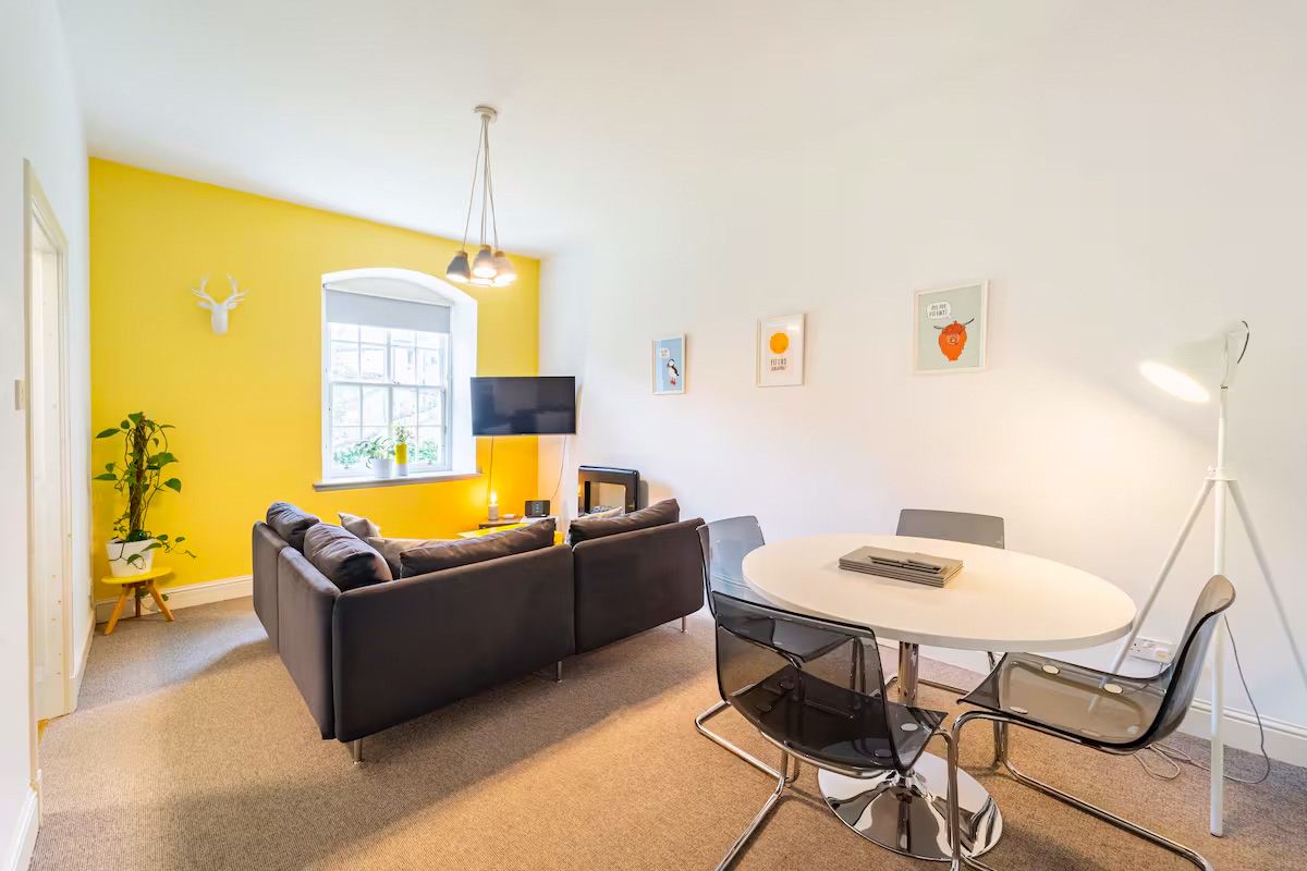 Stylish open-plan living area with bold yellow accent wall, round dining table, and dark sofa.