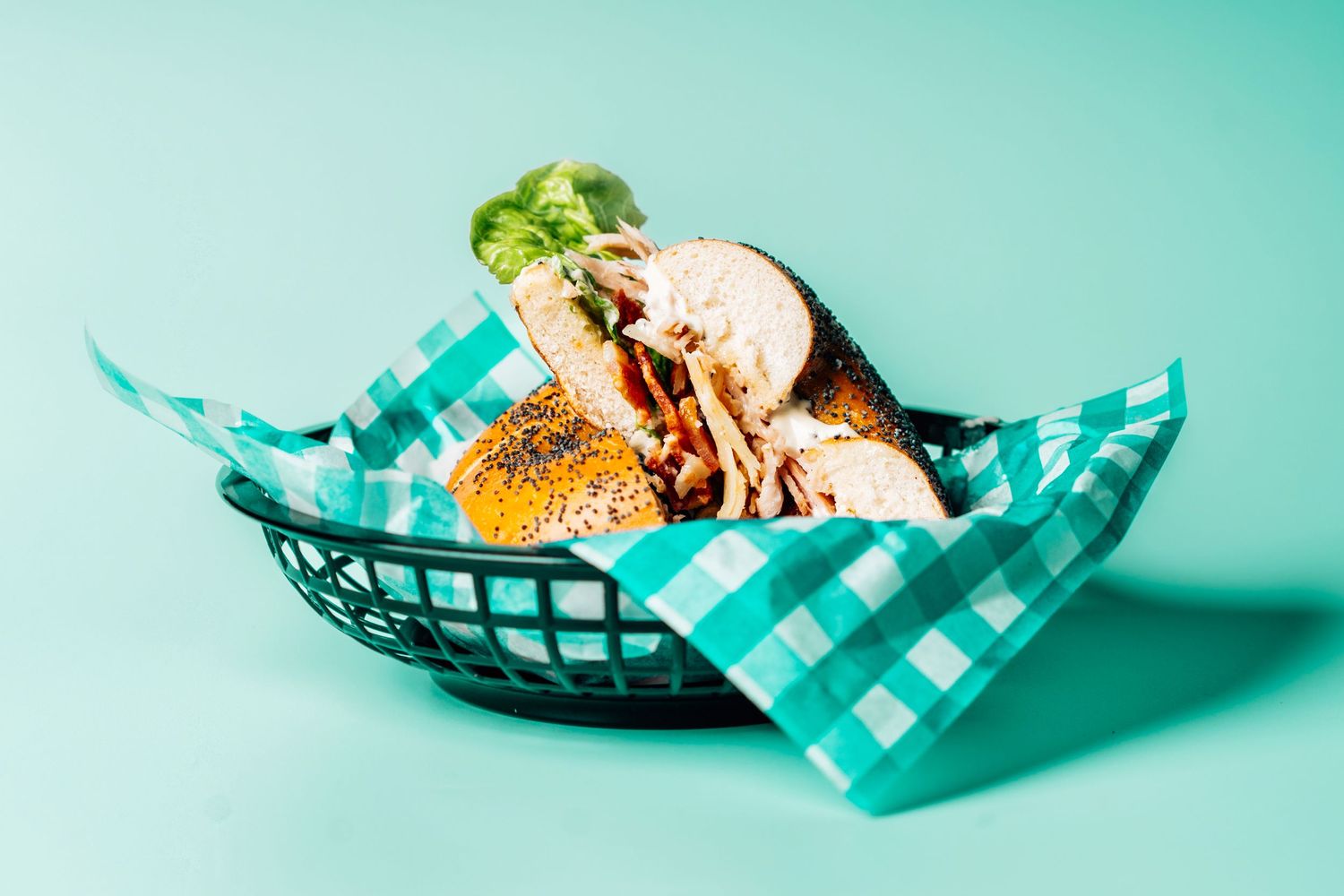 A toasted sesame bagel sandwich served in a black basket with teal checkered paper on a mint background.