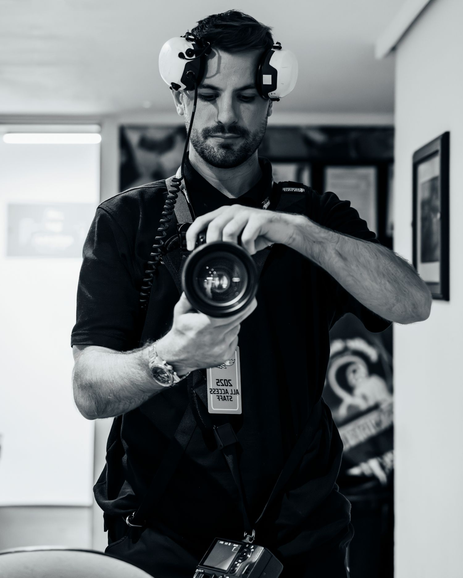 Black and white photo of a bearded male photographer holding a DSLR camera, wearing headphones and a press badge.
