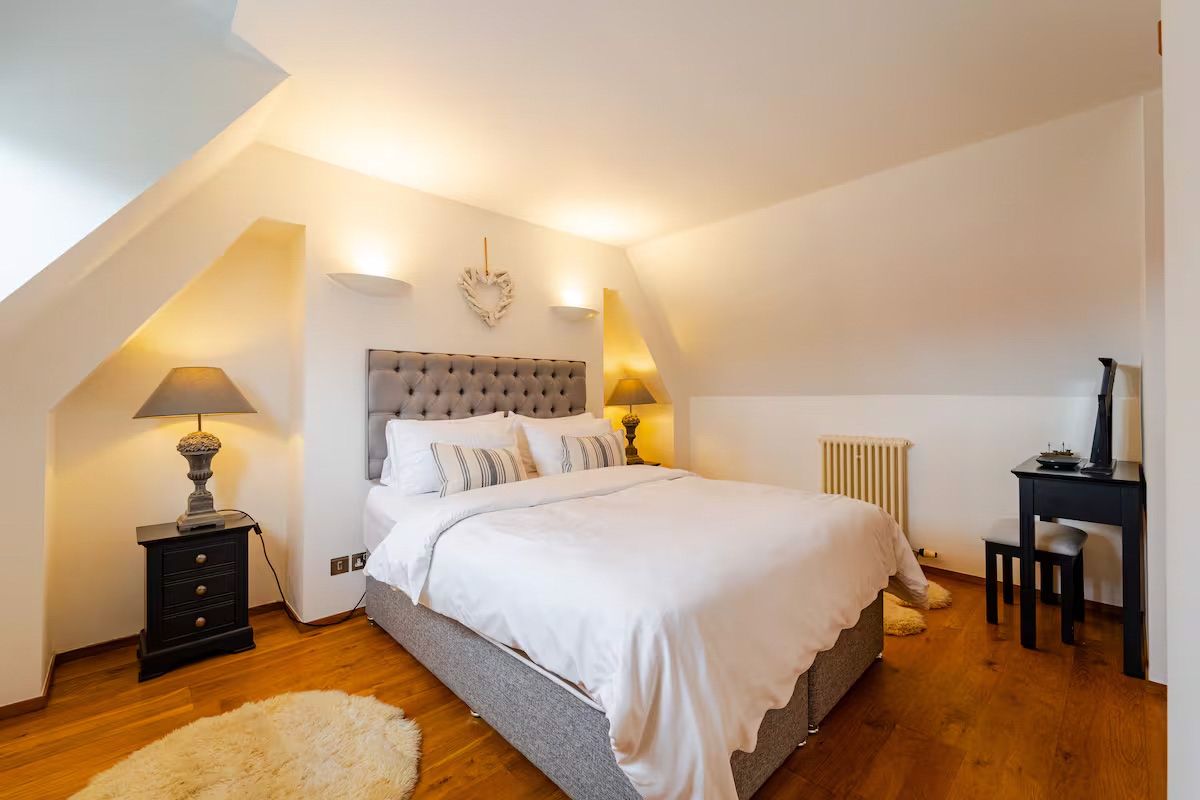 Elegant attic bedroom with upholstered headboard, white bedding, warm lighting, and wooden floor.