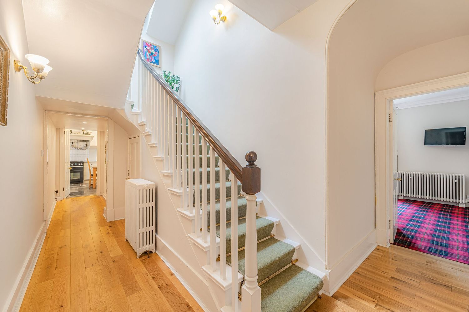 Classic white staircase with wooden banister, light hardwood floors, radiator, and glimpse of tartan carpet room.