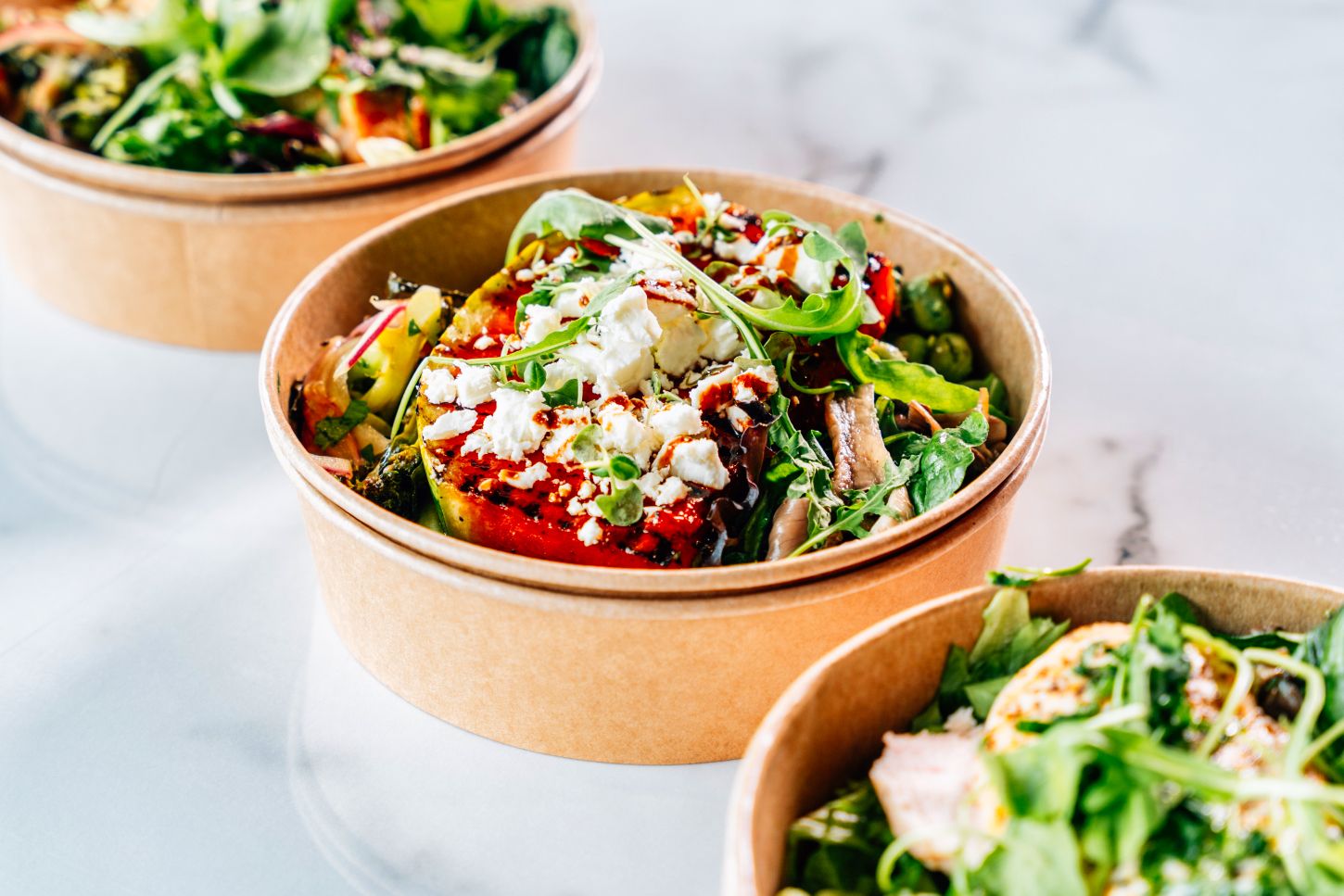 Fresh grain bowls with roasted vegetables, greens, and toppings in eco-friendly kraft paper cups.