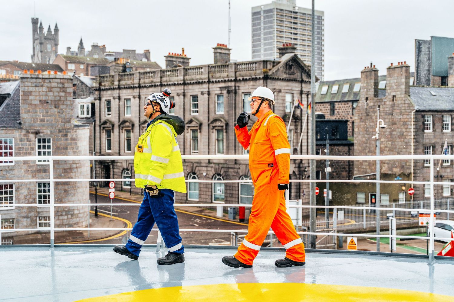 Two marine workers in hi-vis safety gear walk across a ship helipad in Aberdeen port.