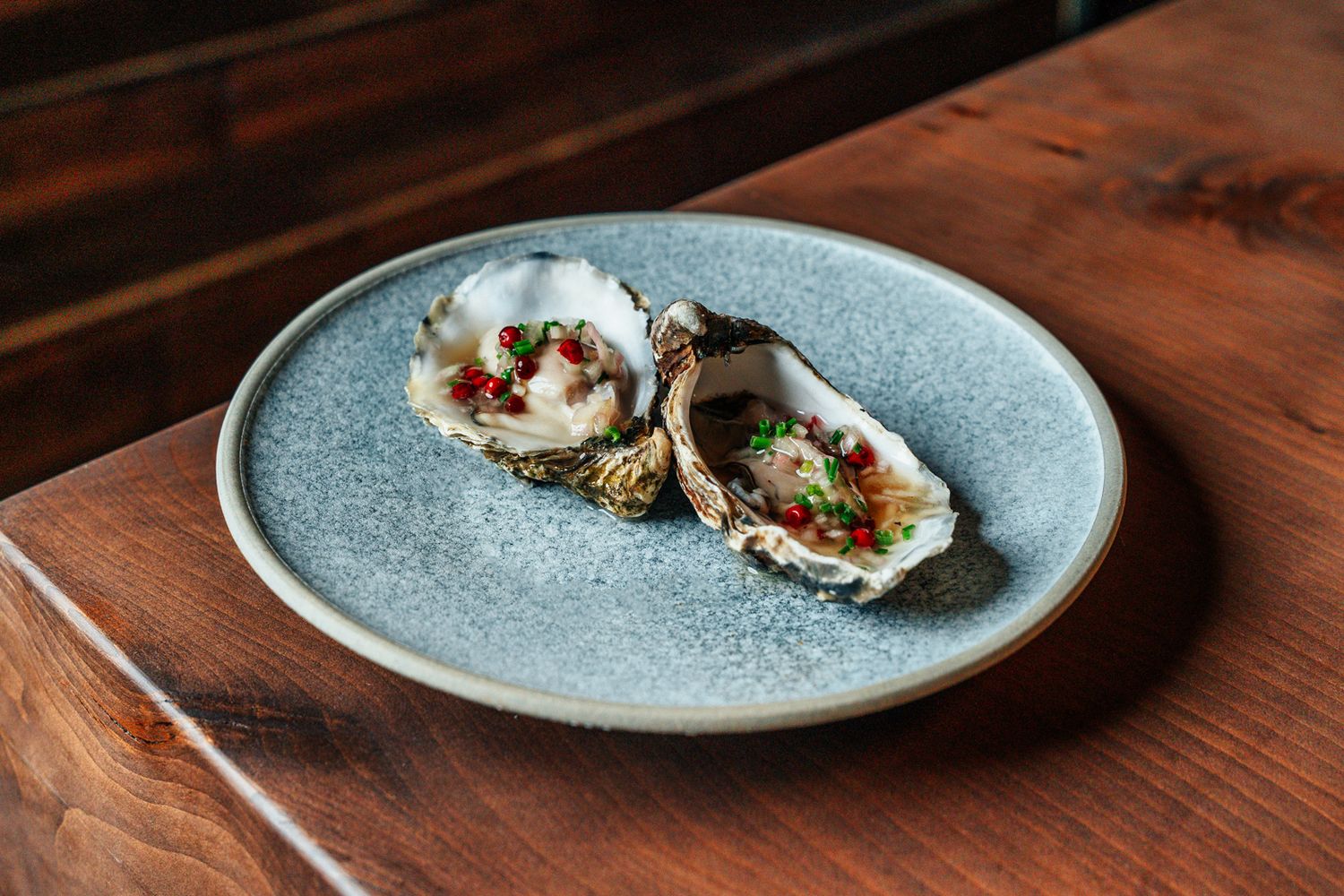 Two fresh oysters on the half shell topped with diced red and green garnish on a grey ceramic plate.