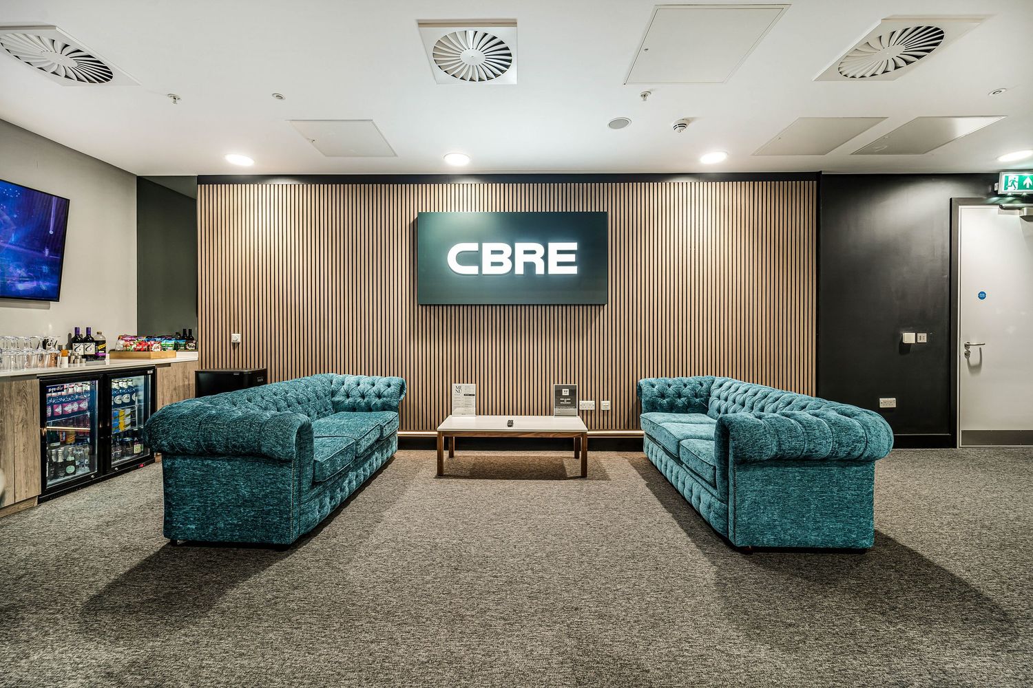 CBRE branded lounge area with two teal Chesterfield sofas, coffee table, and illuminated company signage.