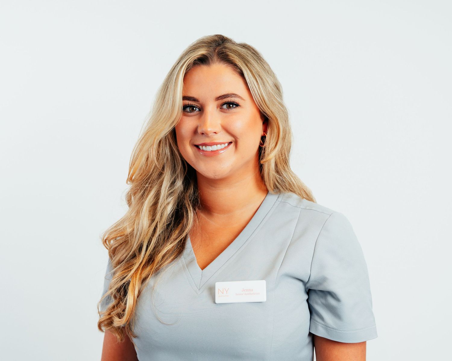 Young blonde woman in gray medical scrubs with name badge smiling confidently against white background.