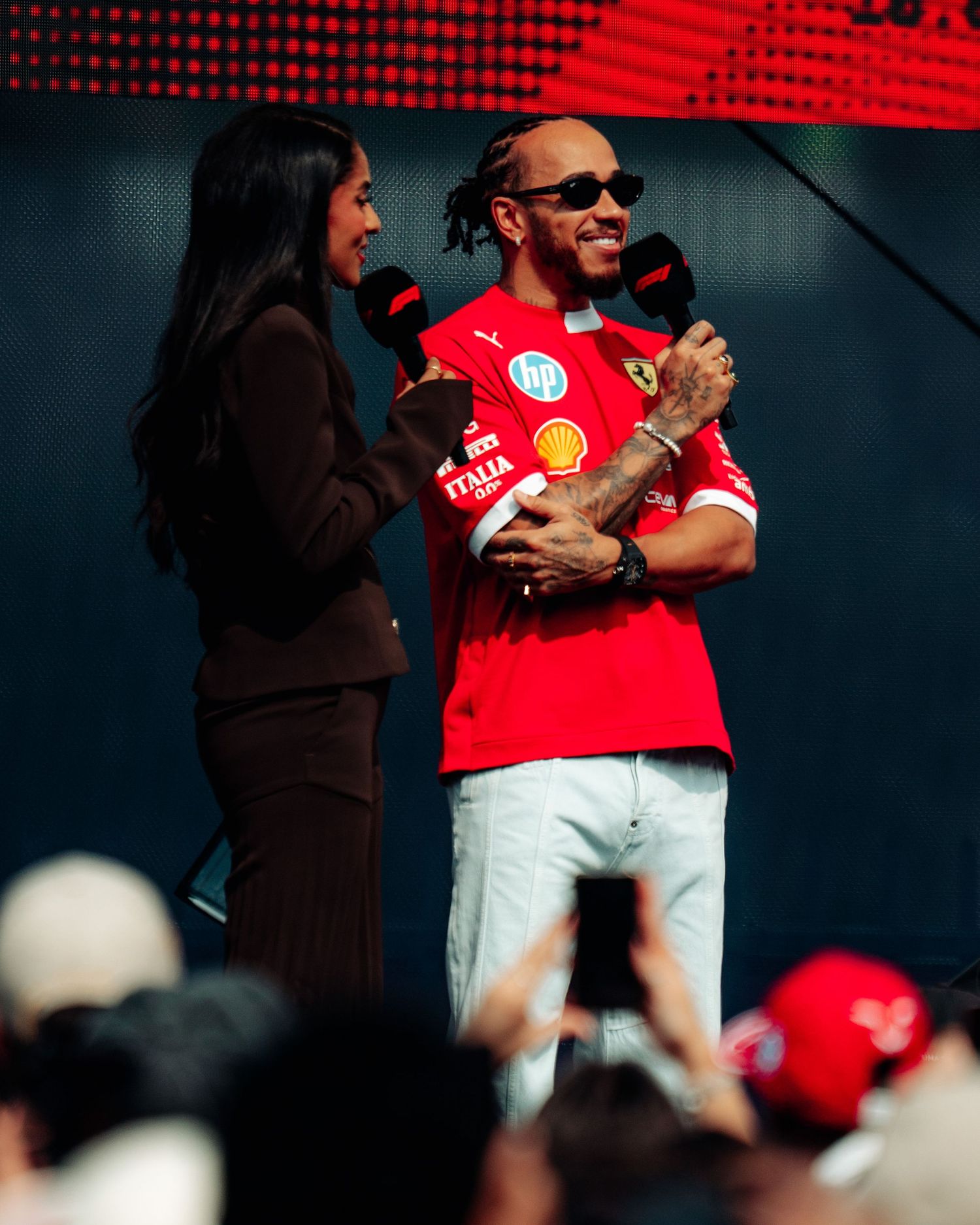 Lewis Hamilton in Ferrari shirt smiles during an on-stage interview at a Formula 1 fan event.
