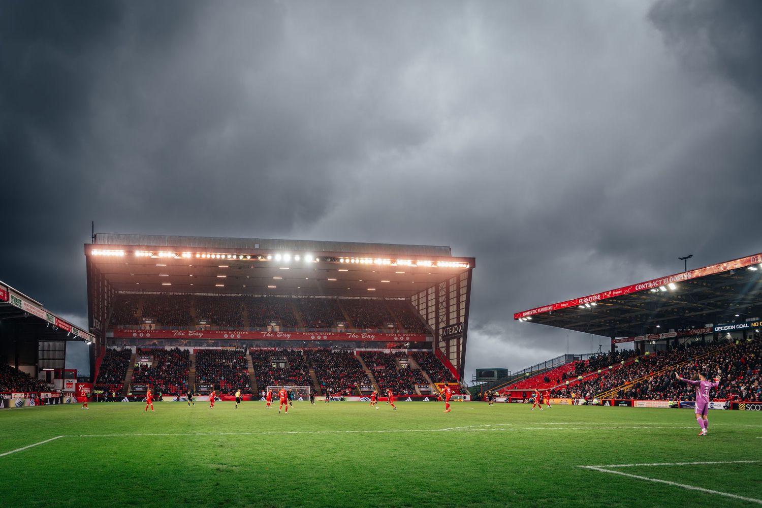 Panoramic view of a football stadium with players on pitch, bright floodlights, and dramatic grey storm clouds.