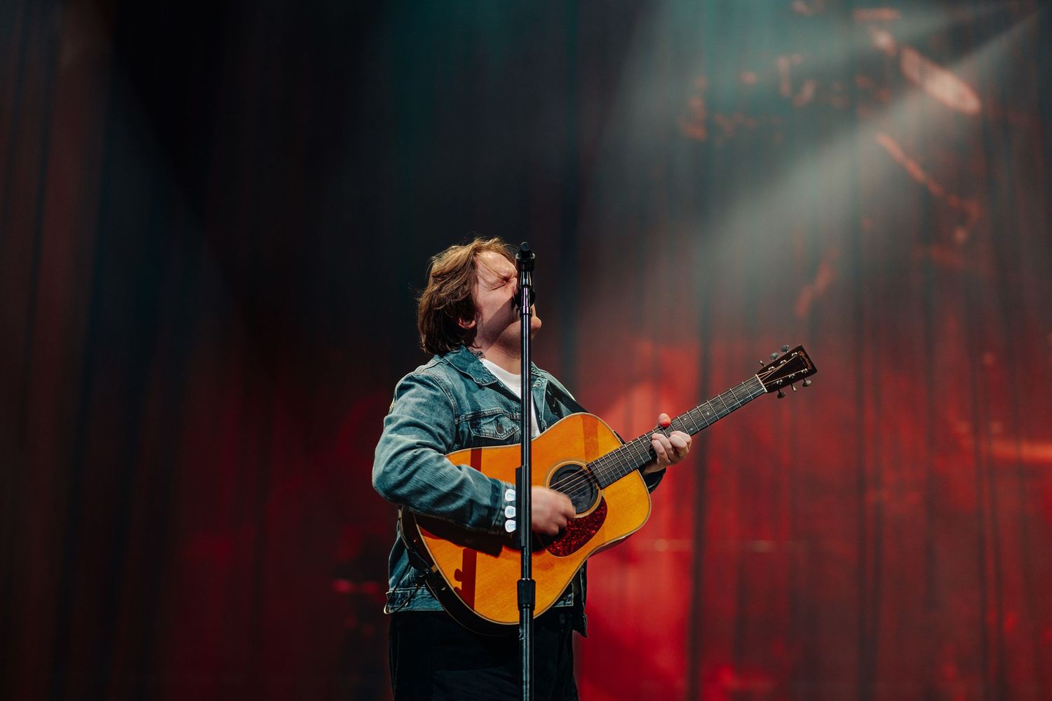 Male guitarist singing on stage with dramatic red and white spotlights behind him.