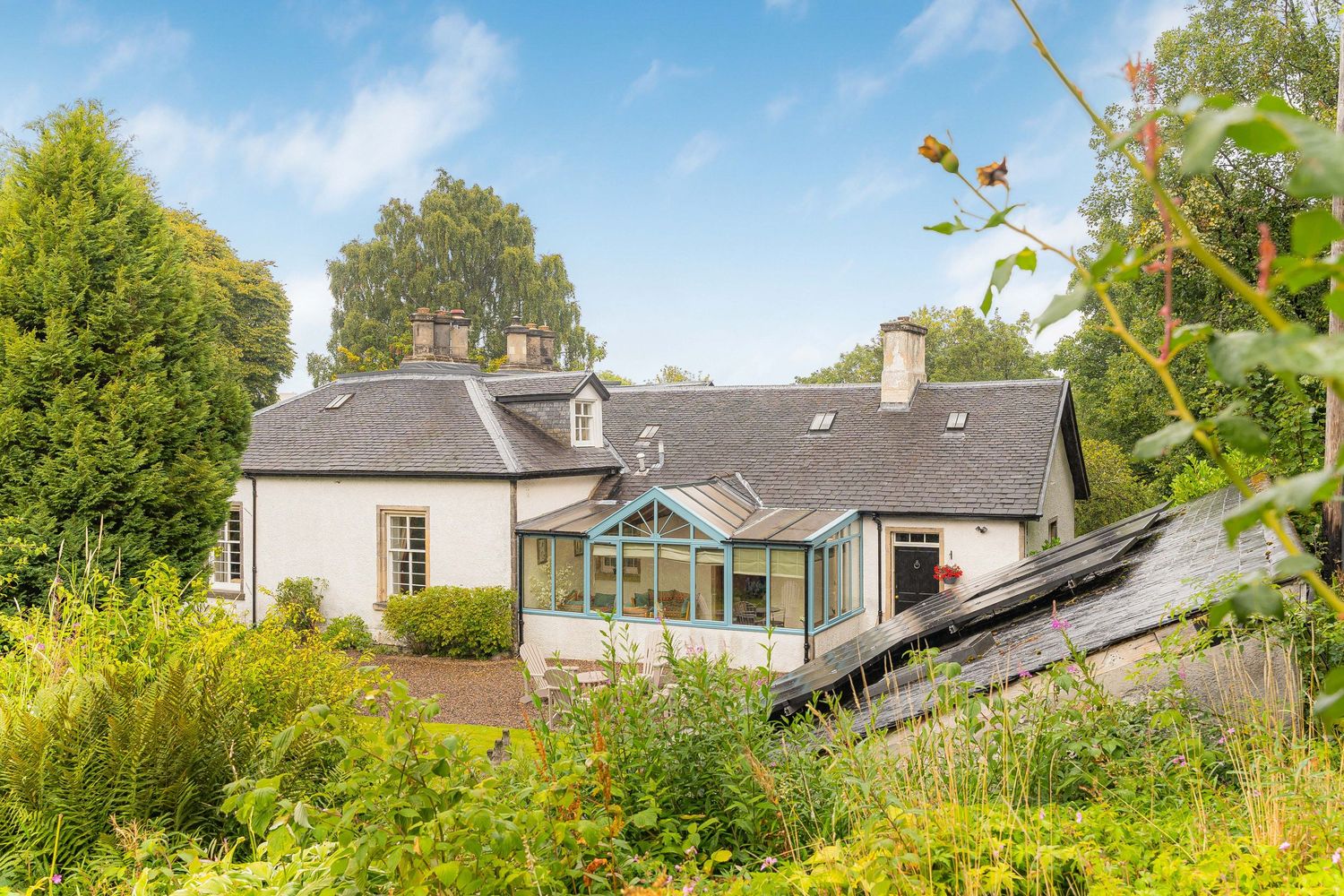 Traditional white Scottish cottage with slate roof, conservatory extension, surrounded by wild garden and mature trees.