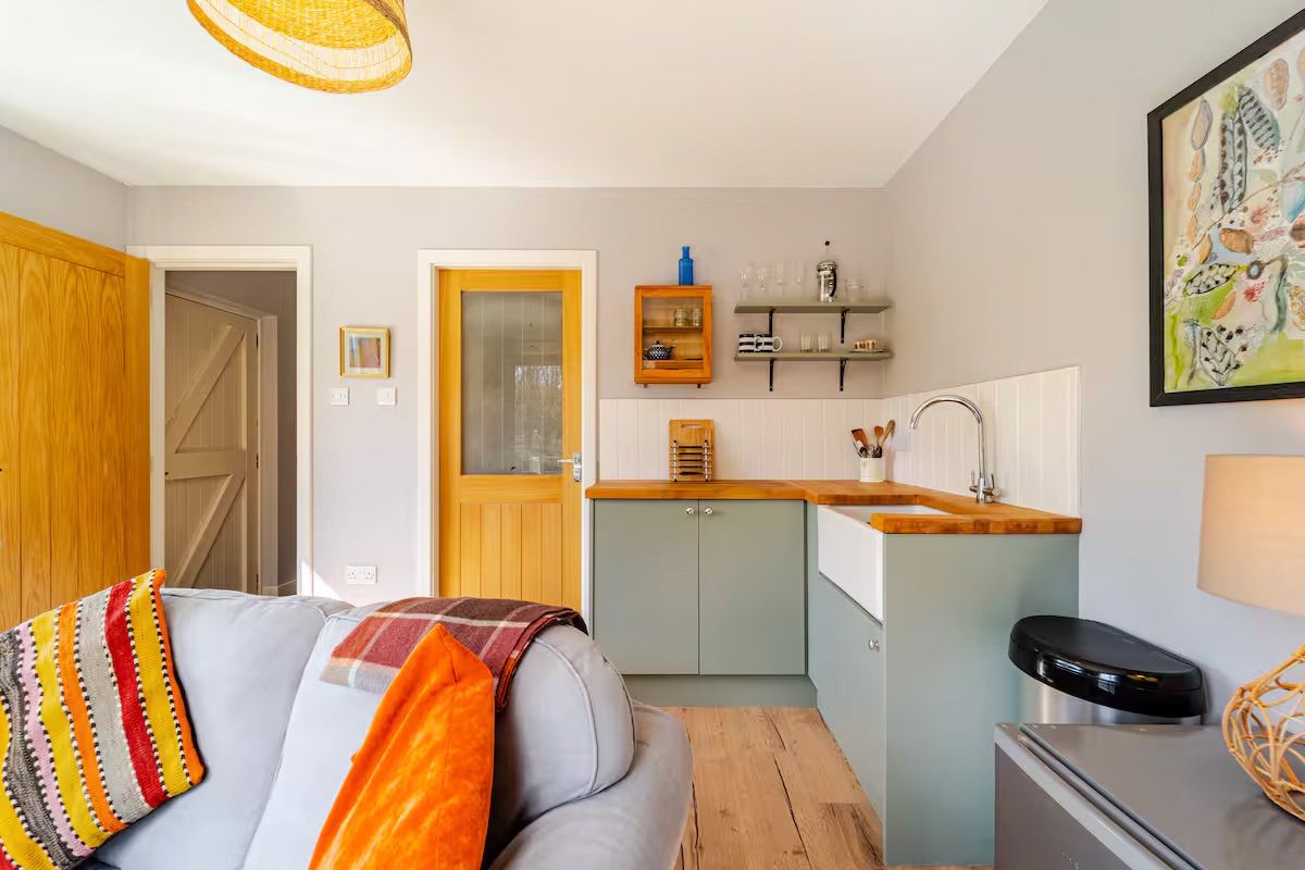 Cozy studio apartment with sage green kitchen, wooden countertop, Belfast sink, and vibrant orange accents on sofa and door.