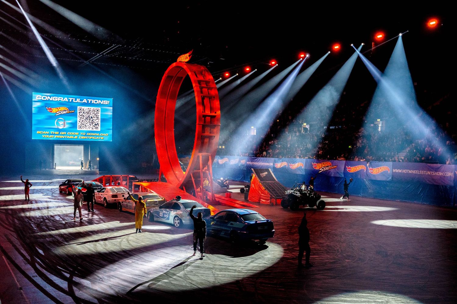 Cars and performers surround a glowing red loop at a Hot Wheels arena show finale with stage lighting.